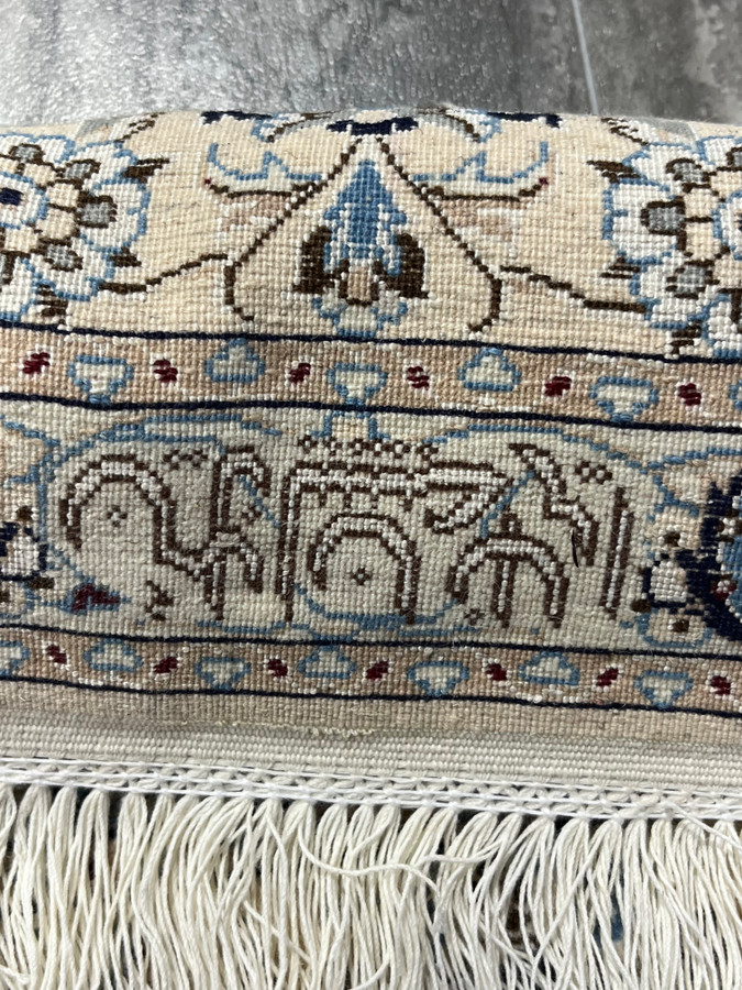 Close-up of Persian Nain 6LA Habibian signature woven into the border, showing fine wool and silk details, ivory ground, soft blue floral motifs, and hand-finished fringe