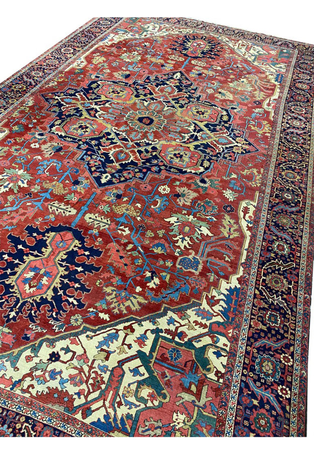 Angled view of oversized antique Heriz mansion rug showing soft tomato red field and ivory spandrels