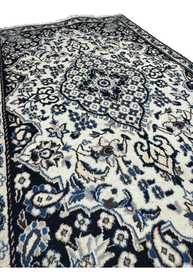 Angled studio view of Persian Nain rug from the foot end, layered floral medallion glowing against ivory and navy palette