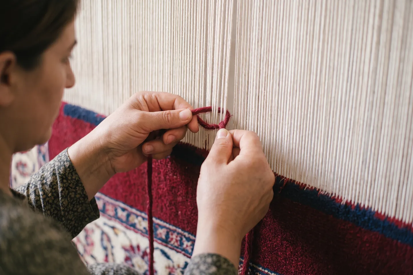 A weaver's hands looping a strand of red wool around two cotton warp threads on a vertical loom