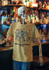 Skeleton mannequin at a lively bar scene wearing the Brew Good Bones – Dancing Skeletons Tee by Coo Coo’s Coffee in Mustard; colorful skeletons dance with coffee beans, adding a fun and spooky twist to coffee culture fashion.