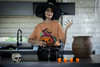 Woman dressed as a witch stirring a cauldron while wearing the Jack-O-Latte – Pumpkin Spice Brew Tee by Coo Coo’s Coffee — festive Halloween pumpkin latte design with bats and coffee vibes.