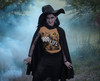 Smiling witch in black cape and hat standing in fog while wearing a yam orange “Boo Life” Halloween graphic tee with pumpkins, bats, and haunted house – Coo Coo’s Coffee spooky season shirt.
