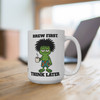 Brew First Think Later 15oz mug — a frazzled, wild-haired Bert the gecko in sandals and floral shorts, droopy-eyed and clutching a steaming coffee cup, with "Brew First." above and "Think Later." below in bold black lettering, plus a Coo Coo's Coffee mark on the side — Coo Coo's Coffee Brew Good Vibes funny morning mug.