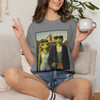 The Americano Gothic coffee tee — two geckos posed as the couple from Grant Wood's American Gothic in front of a white gothic-window farmhouse; Bert in sunglasses holding a pitchfork, Gertie in pearls beside him, with "The Americano Gothic" and a red Florida Brew Crew line beneath — Coo Coo's Coffee Brew Good Vibes garment-dyed Comfort Colors art parody tee.