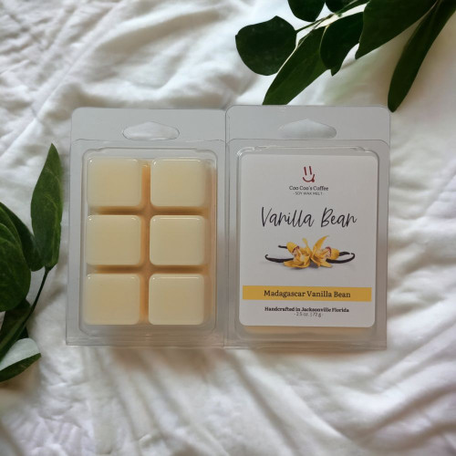 Vanilla Bean wax melt clamshell on white blanket with green leaves, cozy flatlay by Coo Coo’s Coffee