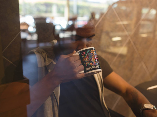 Person sipping from Wake the Art Up neon coffee mug while working in a café behind a window