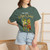 Close-up of the cartoon gecko Bert playing cowbell on a Havana coffee tour graphic print on the More Coffee, More Cowbell t-shirt, blue spruce