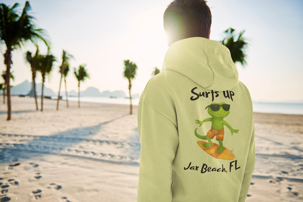 Back view of a man on the sand wearing the Bert’s Surf Club – Jax Beach Hoodie by Coo Coo’s Coffee, featuring Bert the gecko surfing graphic with ‘Surf’s Up – Jax Beach, FL’ text.