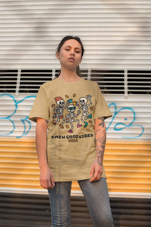 Young woman standing confidently in front of a graffiti wall wearing the Brew Good Bones – Dancing Skeletons Tee by Coo Coo’s Coffee in Mustard; playful skeleton characters dancing with coffee beans add a bold streetwear vibe.