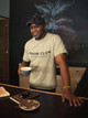 Tan Brew Club – Small Batch Society Tee by Coo Coo’s Coffee, modeled at café counter with cup in hand — clean unisex coffee shirt for Small Batch of Friends community.