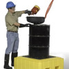A worker in protective gear is pouring liquid from a red container into a black drum positioned on a yellow spill pallet without a drain.