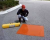 A square orange drain seal displayed flat on a paved surface next to a yellow emergency container.