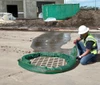 Construction worker in safety gear installing a green drain sock around a storm drain grate at a construction site to prevent debris and runoff from entering.