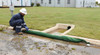 Worker in safety clothing positioning a long green drain sock across a curb inlet to control water flow and capture pollutants.