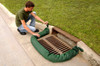 A man kneeling by a curb storm drain, positioning a green filter sock barrier around the grate to prevent debris from entering.