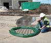 A construction worker wearing a safety vest and helmet kneels on pavement, arranging a green filter sock around a storm drain near a construction site.