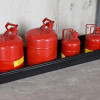 Four red safety cans of varying sizes placed in a black utility tray for spill containment, displayed against a concrete wall.