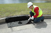 Worker in safety gear placing a black drain guard with a built-in filter over a curb storm drain near a grassy area.