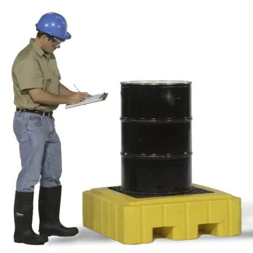 A worker wearing safety gear, including a blue helmet and black boots, is inspecting a black drum placed on a yellow spill pallet while writing on a clipboard.