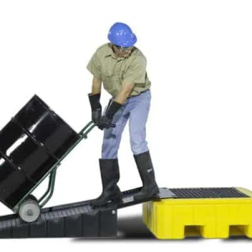 A worker in safety gear is rolling a black drum up a ramp and onto a yellow spill pallet using a hand truck.