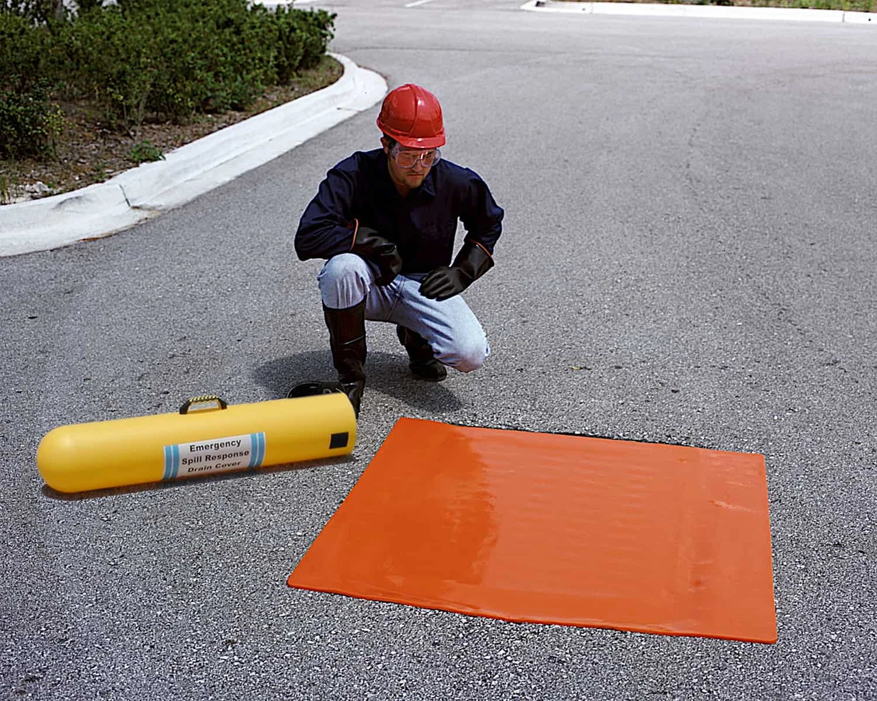 A square orange drain seal displayed flat on a paved surface next to a yellow emergency container.