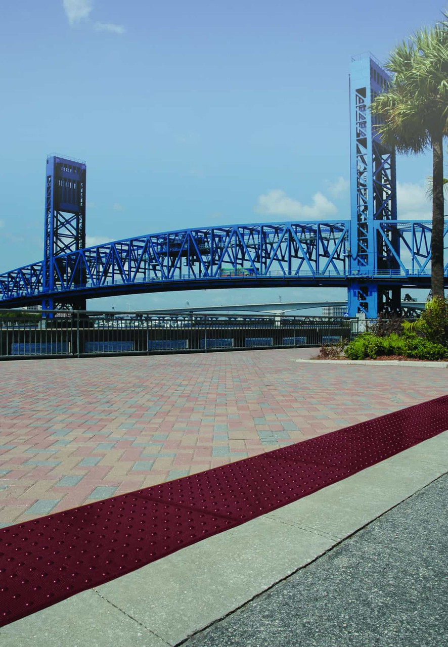 Red tactile ADA pad placed along a sidewalk near a scenic blue bridge.