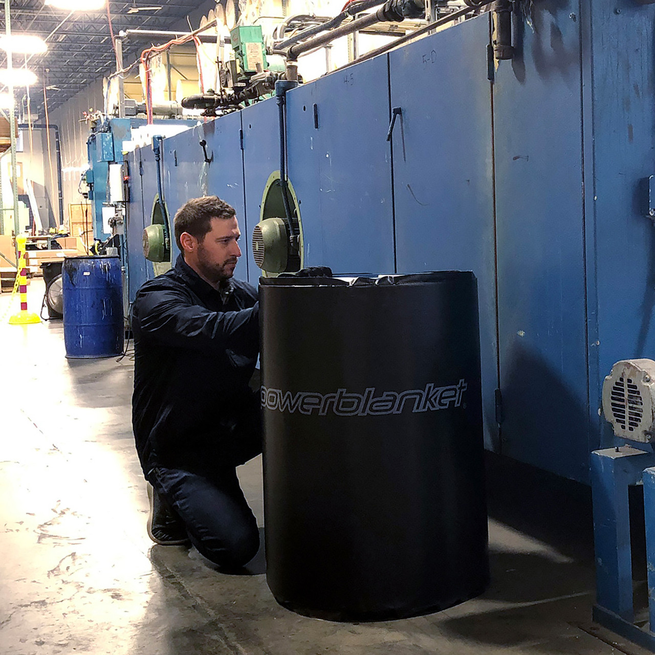 Man kneeling and adjusting a black Powerblanket drum heater around a drum in an industrial facility.