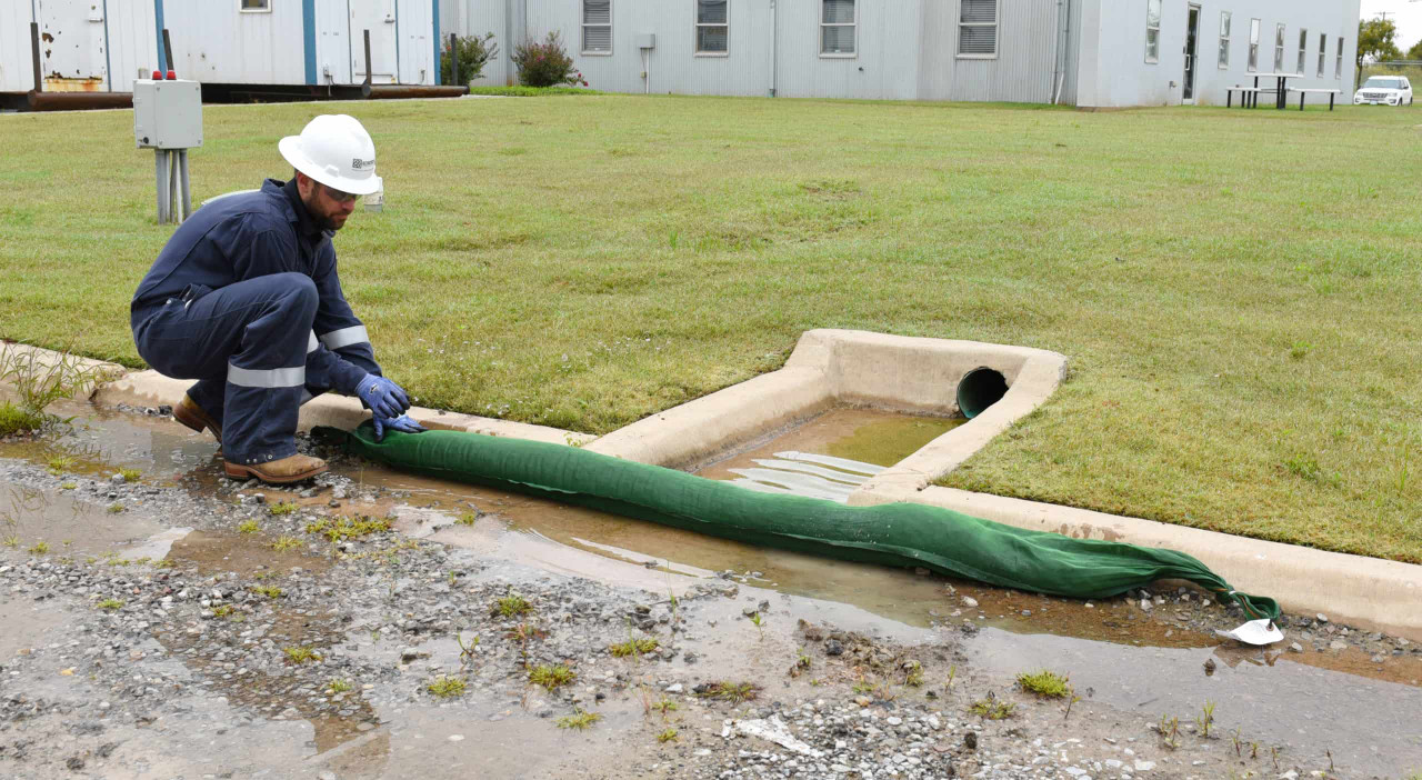 Worker in safety clothing positioning a long green drain sock across a curb inlet to control water flow and capture pollutants.