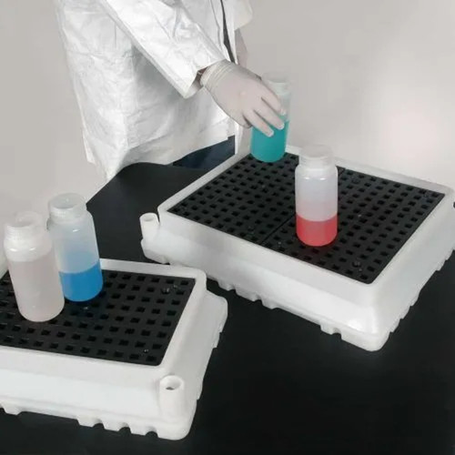 Two white spill trays with black grates holding bottles of clear, blue, and red liquids, used for spill containment in a lab.
