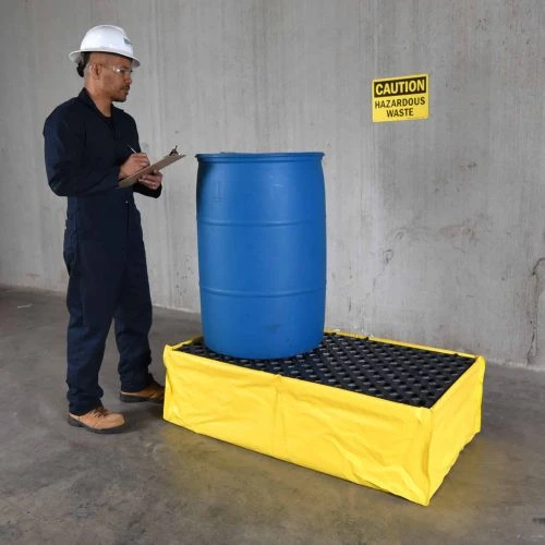 A worker in a white hard hat inspects a blue drum placed on a yellow flexible spill pallet, with a hazardous waste caution sign on the wall.
