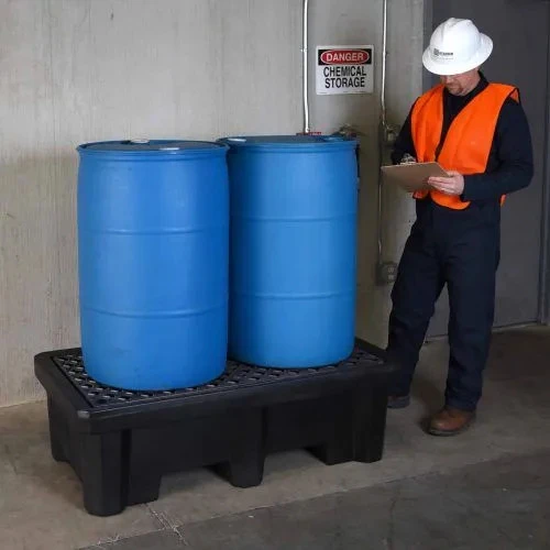 A worker in a hard hat and safety vest inspecting two blue drums placed on a black spill containment pallet in a chemical storage area.