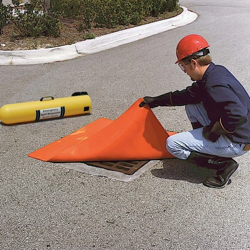 A worker in safety gear lifting the edge of an orange drain seal while covering a metal drain grate on a roadway.