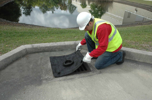 Worker wearing a hard hat, safety vest, and gloves installing a black drain cover over a storm drain inlet on a roadside curb.
