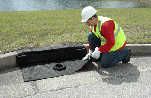 Worker in safety gear placing a black drain guard with a built-in filter over a curb storm drain near a grassy area.