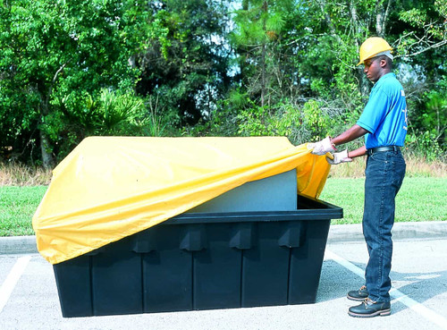 A worker covering a tank in a black containment sump with a yellow tarp outdoors.