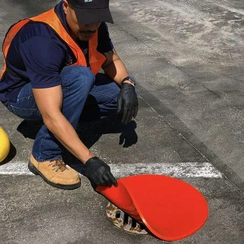 A worker in safety gear lifting the edge of an orange circle drain seal while covering a metal drain grate on a roadway.