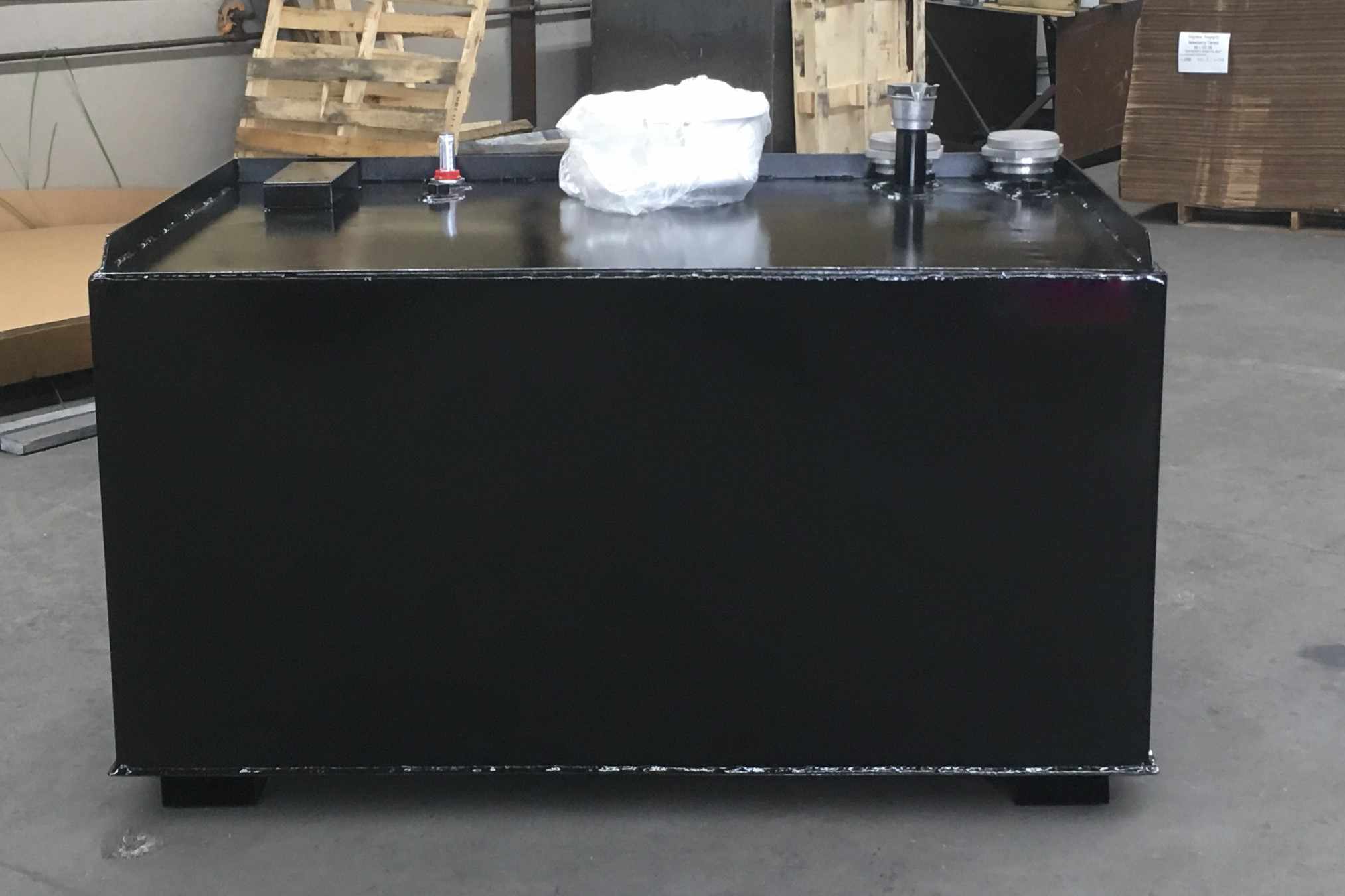 Black rectangular industrial storage tank with multiple fittings on top, positioned indoors on a concrete floor.