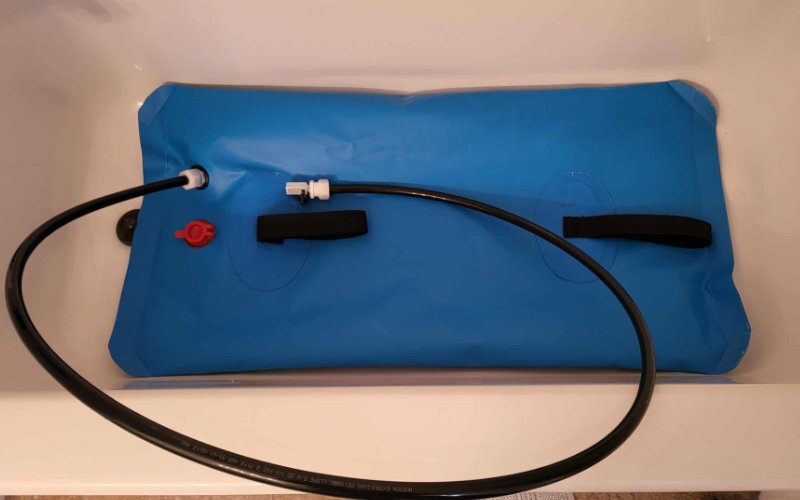 Small blue flexible water bladder with attached hoses and fittings, designed for compact water storage, positioned in a bathtub.