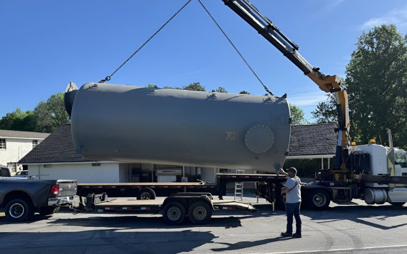 Large gray fiberglass tank being lifted by a crane onto a trailer, ready for transport to an oil and gas storage site.