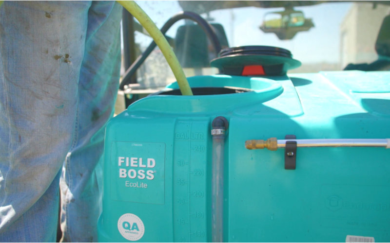 Field Boss Ecolite turquoise tank being filled with a yellow hose, featuring a liquid gauge and QA mark, attached to agricultural equipment.
