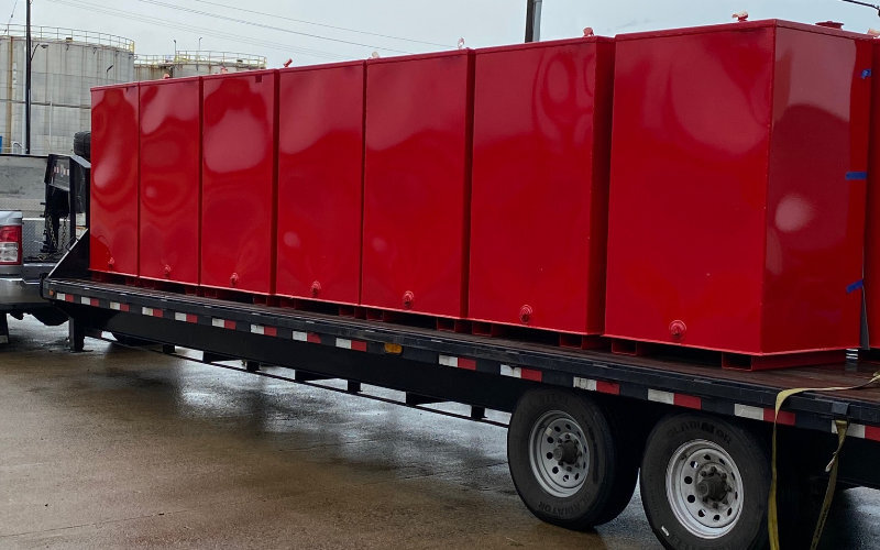 Multiple red steel tanks lined up on a flatbed trailer, ready for transport in a rainy industrial area with storage tanks visible in the background.