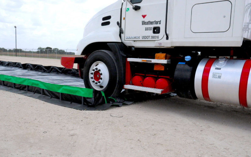 White truck driving over a green and black containment liner setup, demonstrating effective spill containment on a sandy surface.