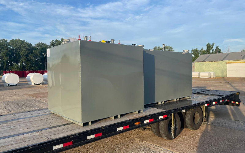 Two large gray rectangular steel tanks secured on a flatbed trailer, situated in an outdoor industrial lot with storage tanks visible in the background.