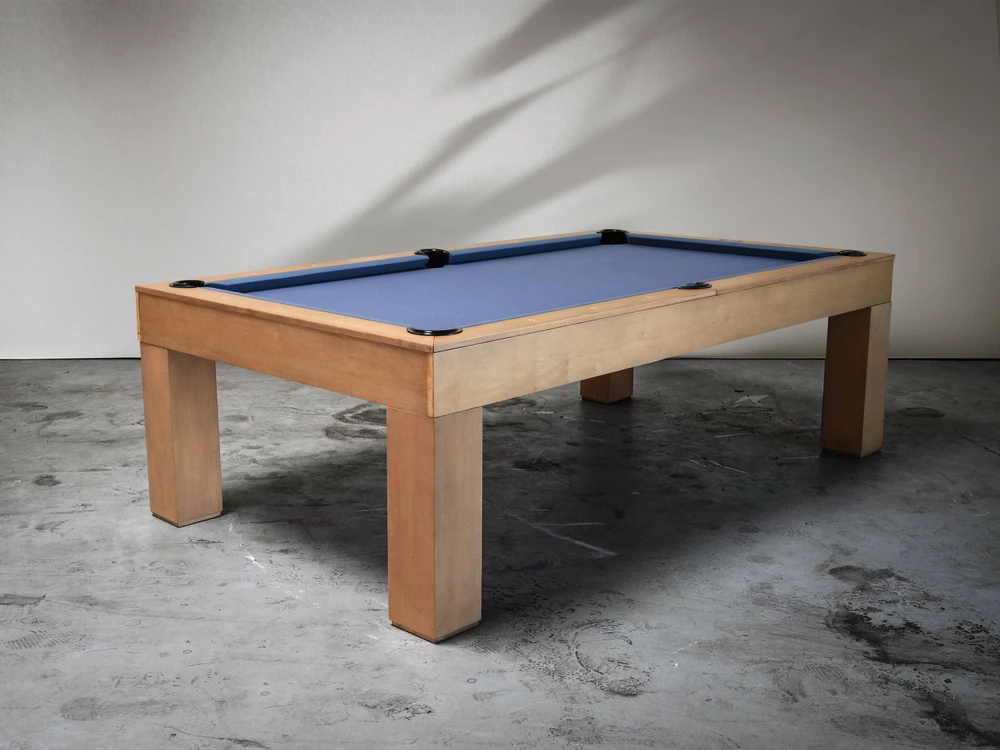 Full 3/4 lifestyle view of the Paula Parsons Slate Pool Table by Doc & Holliday, featured in glazed maple with subtle distressing, staged on smooth concrete flooring with soft natural light and a minimal modern backdrop highlighting clean Parsons legs and timeless craftsmanship