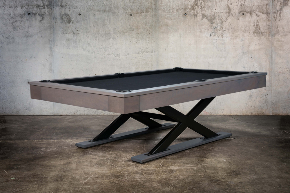 The Metalsmith by Doc & Holliday pool table photographed from the corner angle, highlighting the oversized X-leg steel base and clean hardwood apron. The dark felt and matte finishes echo the feel of a designer loft or urban penthouse game room.