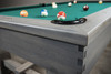 Close-up of the Churchill Pool Table corner in Smoked Maple distressed finish, showing finger-jointed apron and buffalo nickel sight, with blurred concrete wall in the background.