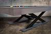 Architectural focus on the Metalsmith’s oversized X-leg steel base, powder-coated in deep black, set against textured flooring. The legs ground the table with an industrial edge reminiscent of sculptural furniture in Restoration Hardware showrooms.