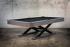 The Metalsmith by Doc & Holliday pool table photographed from the corner angle, highlighting the oversized X-leg steel base and clean hardwood apron. The dark felt and matte finishes echo the feel of a designer loft or urban penthouse game room.