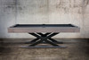 The Metalsmith by Doc & Holliday pool table standing against a raw concrete wall — modern industrial minimalism. Black Championship felt contrasts with the powder-coated steel X-legs, creating a bold architectural silhouette.
